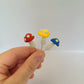 Three mushrooms mounted on metal pins are held against a white background. One is red, one yellow and one blue. The tops are textured with white spots.