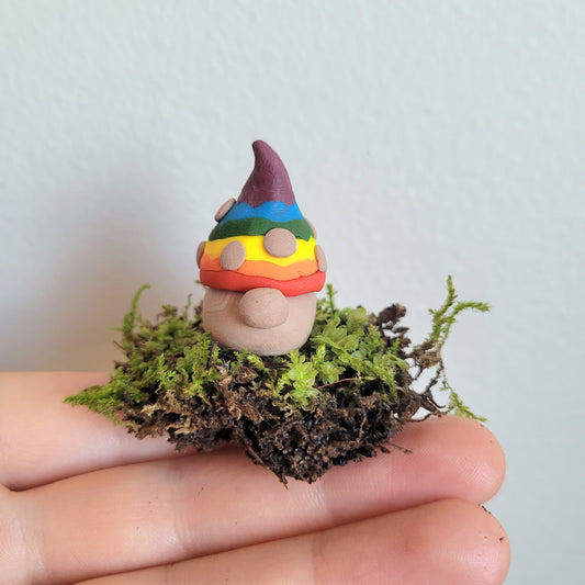 A handmade clay gnome sits on a small patch of green moss. The gnome has white skin with a circular nose and a rainbow striped pointy hat with the colours of the Pride flag. It has textured white spots on its hat.