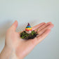 A zoomed out image of a handmade clay gnome sitting on a small patch of green moss held by the artist's hand. The gnome has white skin with a circular nose and a rainbow striped pointy hat with the colours of the Pride flag. It has textured white spots on its hat.