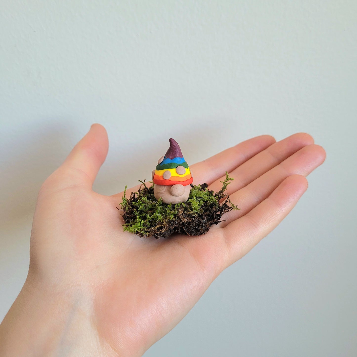 A zoomed out image of a handmade clay gnome sitting on a small patch of green moss held by the artist's hand. The gnome has white skin with a circular nose and a rainbow striped pointy hat with the colours of the Pride flag. It has textured white spots on its hat.