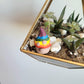 A handmade clay gnome sits in a gold framed glass geometrical terrarium filled with succulents and pebbles. The gnome has white skin with a circular nose and a rainbow striped pointy hat with the colours of the Pride flag. It has textured white spots on its hat.