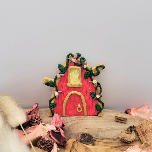 The princess fairy door rests on an oak wood platform surrounded by preserved pink flower petals, wood pieces and willows. It is pink with gold trim, green vines and leaves, and purple flowers.