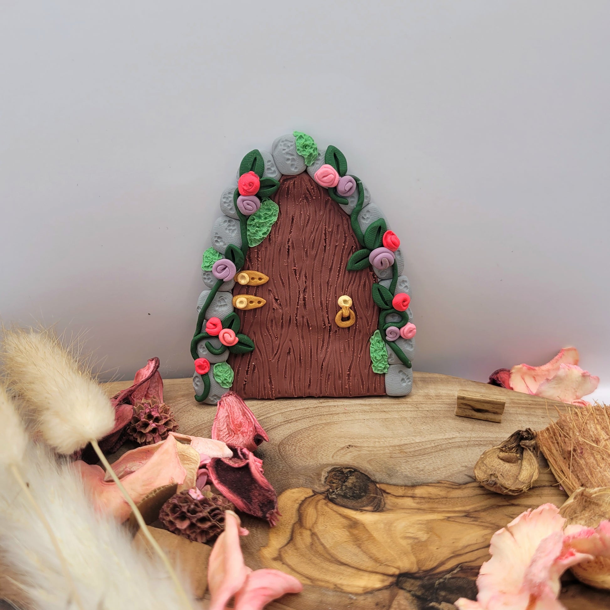 The fairy door rests on an oak wood platform surrounded by preserved pink flower petals, wood pieces and willows. The secret garden door is a maroon wood pattern with grey cobblestone finish and gold hinges and door handle. It is detailed with green vines and pink and purple flowers.