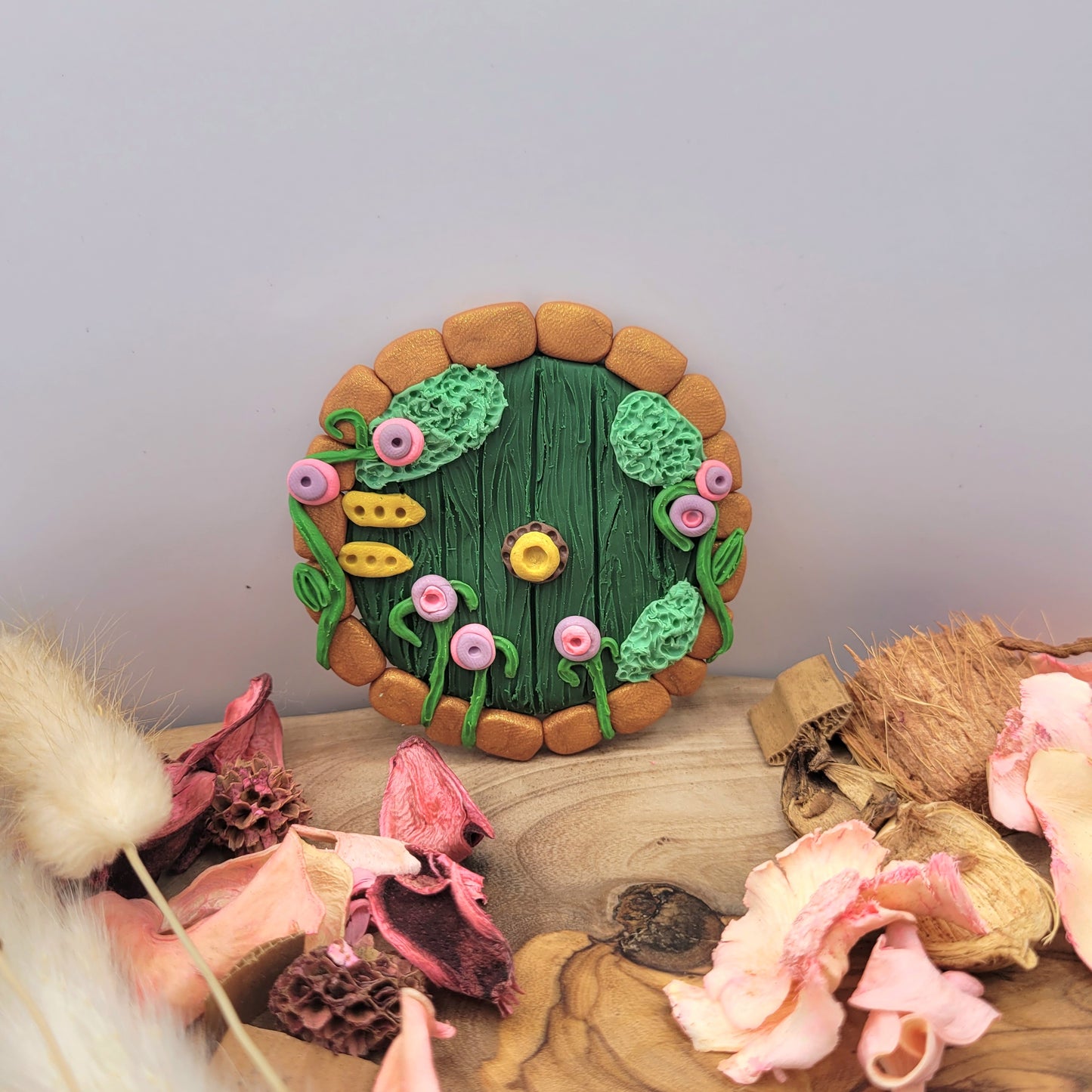 The Hobbit door rests on an oak wood platform surrounded by preserved pink flowers, wood pieces and willows. The Hobbit door is circular and green wood patterned with bronze brick finishes and golden hinges and a doorknob. It is detailed with green grass, moss and bright purple and pink flowers.