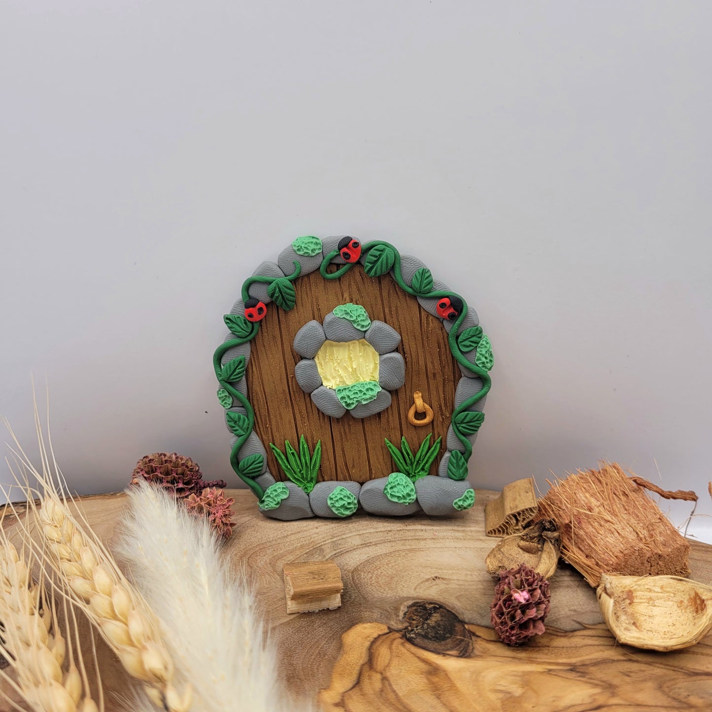 The ladybug fairy door sits on an oak wood platform surrounded by preserved wood pieces and willows. The mini ladybug door is brown wood patterned with grey cobblestone finish, a window and a door knob. It is detailed with green grass, moss, vines and, of course, ladybugs!