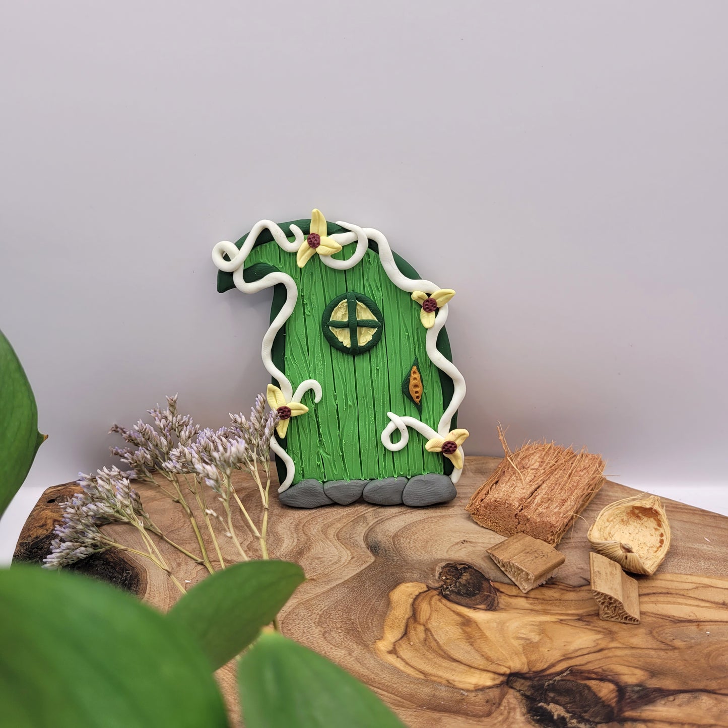 The Elvish fairy door sits on an oak wood platform surrounded by dried flowers and wood. Shaped like a leaf, this Elvish door has green wood patterning, white vine finish, and a window. It is detailed with yellow flowers.