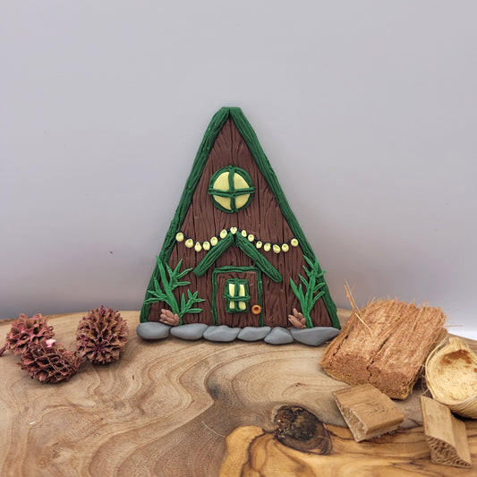The alpine cabin fairy door rests on an oak wood platform surrounded by preserved wood pieces and pinecones. It is brown with a forest green roof and detailed with a window, fairy lights, door, spruce sprigs and pinecones.