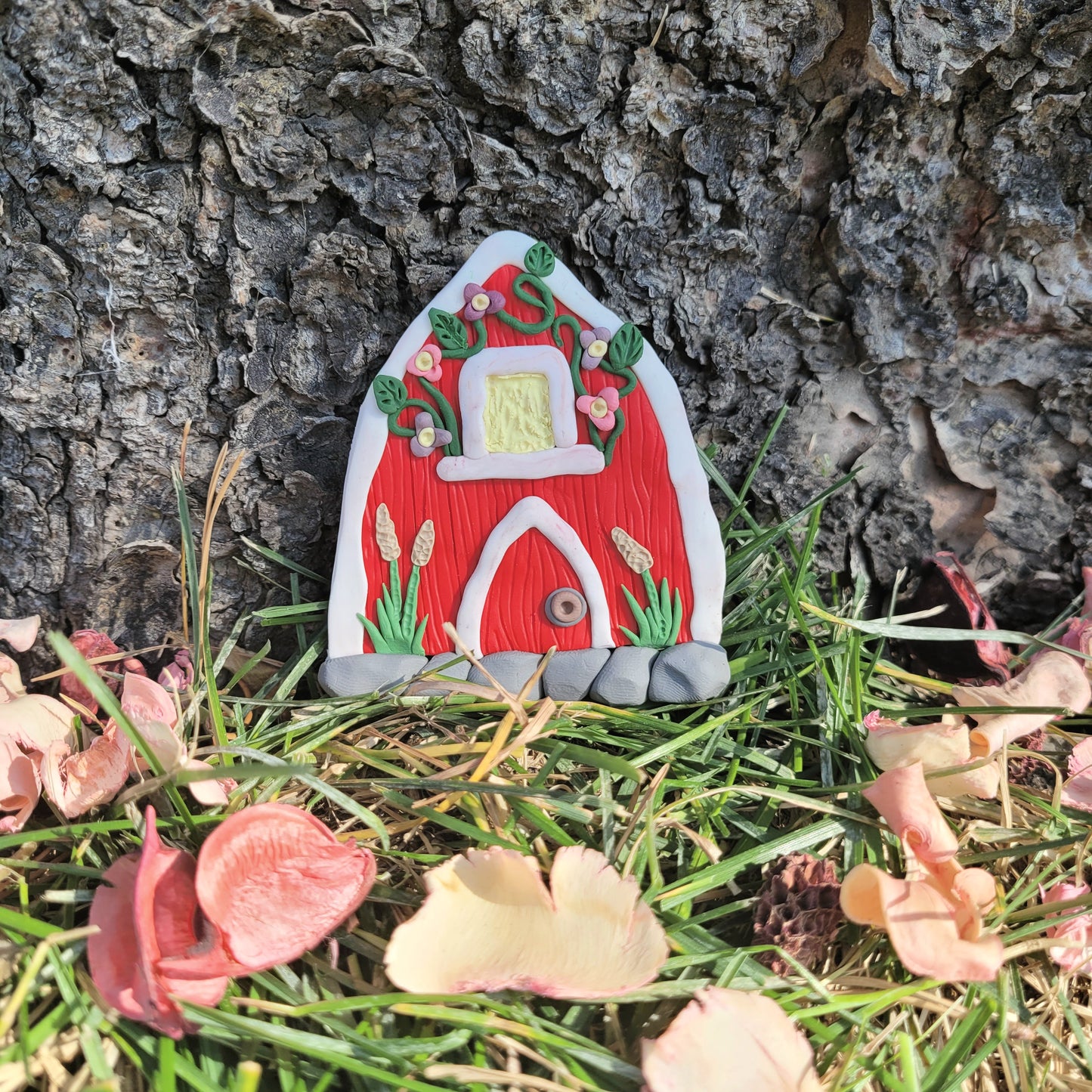 The barn door rests against the bark of a tree surrounded by green grass and pink petals. The barn door is a red wood pattern with white finish, a window, a door, and a cobblestone patterned bottom. It is detailed with pink and purple flowers, grass and some wheat stalks.