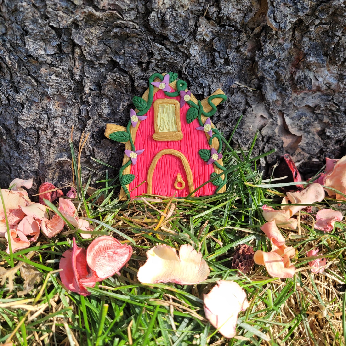 The princess fairy door rests against the bark of a tree outside surrounded by green grass and pink flower petals. The princess fairy door rests on an oak wood platform surrounded by preserved pink flower petals, wIt is pink with gold trim, green vines and leaves, and purple flowers.