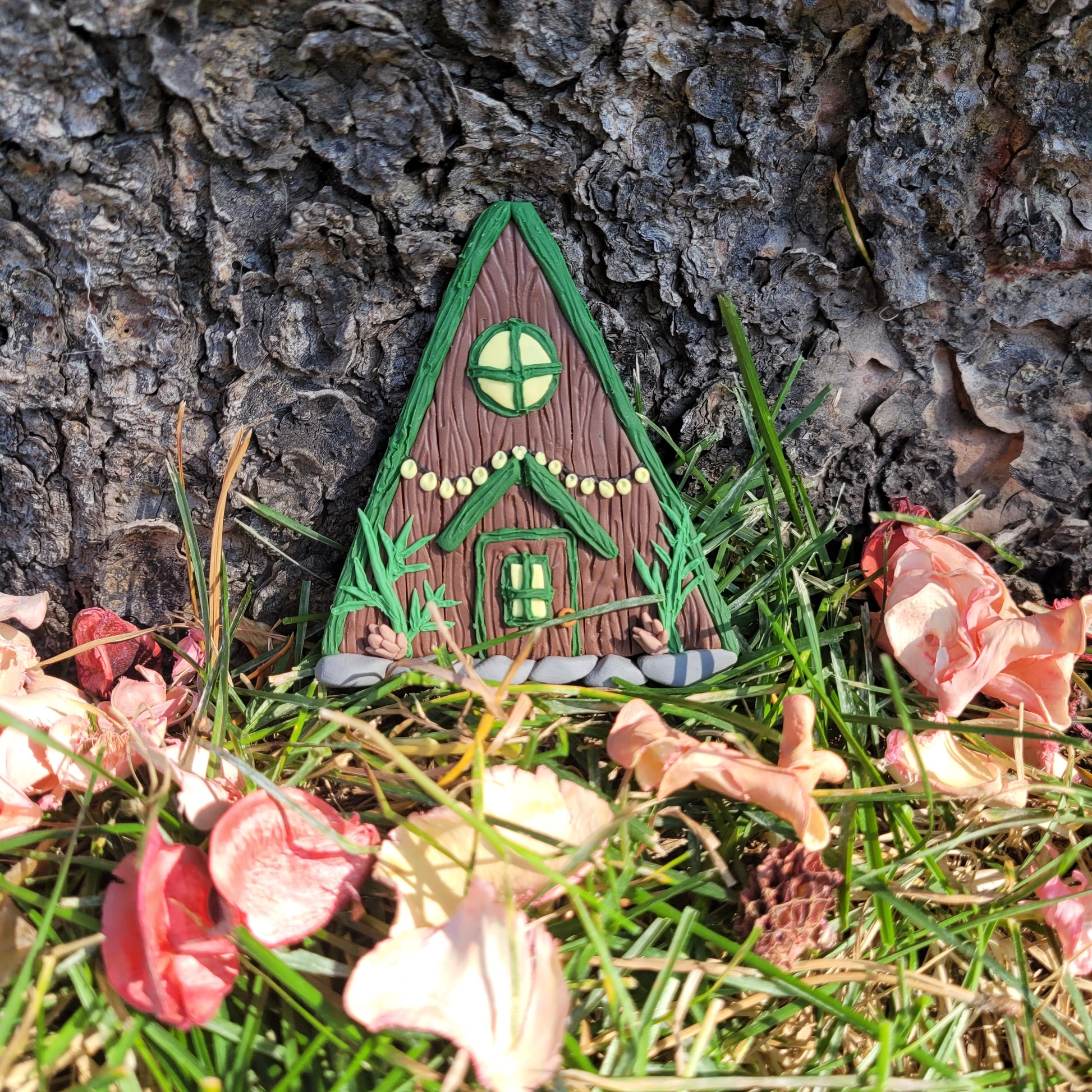 The alpine cabin fairy door rests against the bark of a tree outside surrounded by green grass and pink flower petals. It is brown with a forest green roof and detailed with a window, fairy lights, door, spruce sprigs and pinecones.