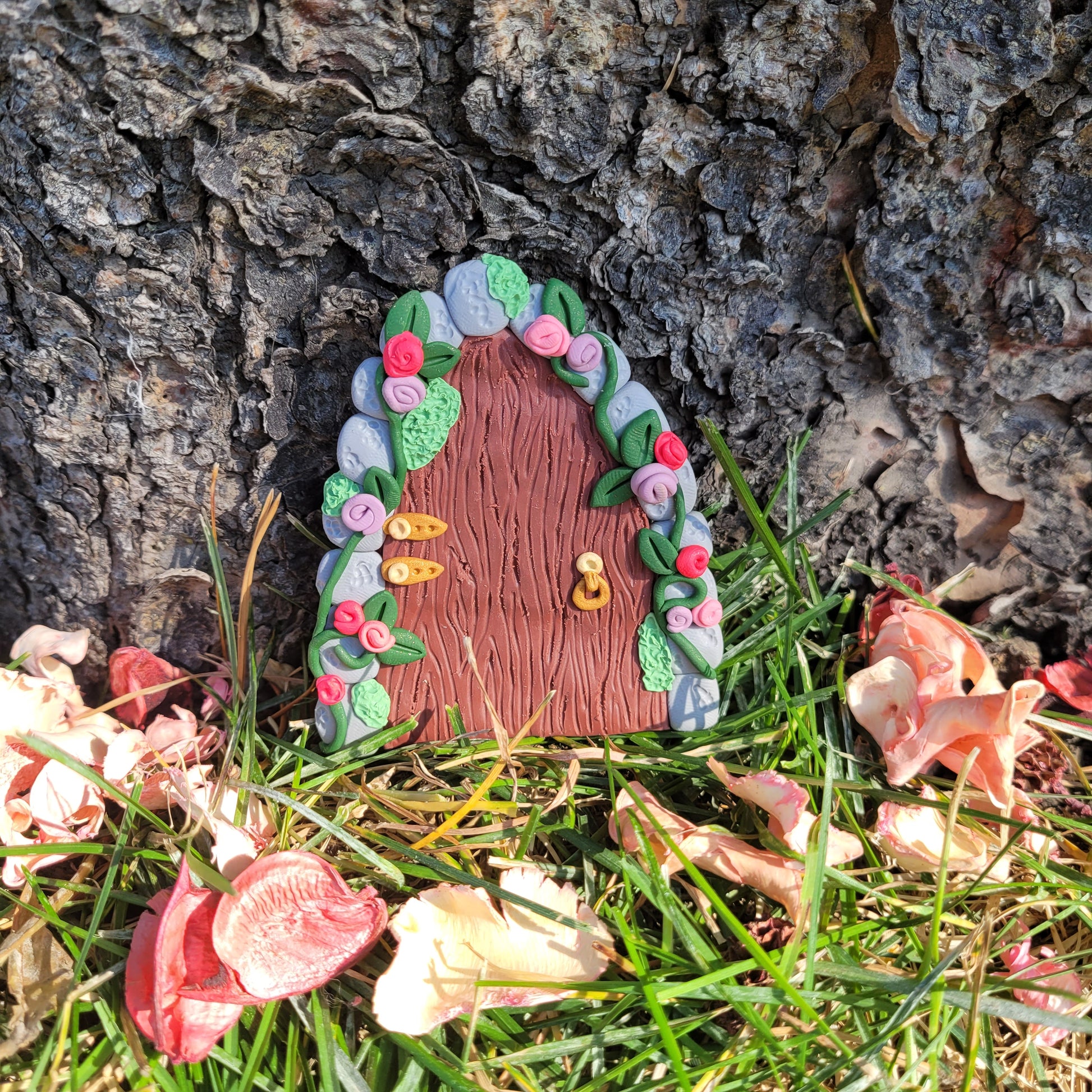 The fairy door rests against the bark of a tree outside surrounded by green grass and pink flower petals. The secret garden door is a maroon wood pattern with grey cobblestone finish and gold hinges and door handle. It is detailed with green vines and pink and purple flowers.