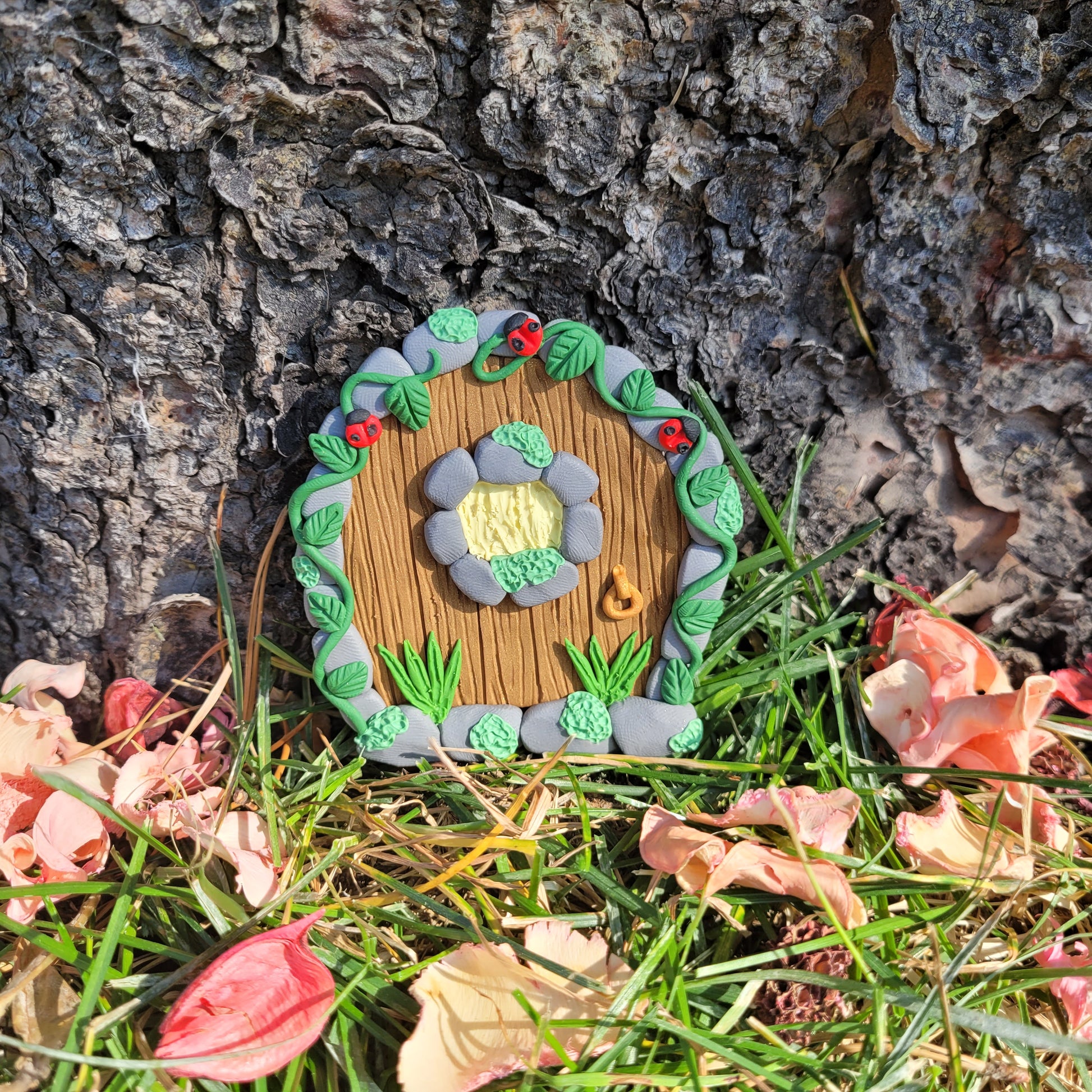 The ladybug fairy door rests against the bark of a tree outside surrounded by green grass and pink flower petals. The mini ladybug door is brown wood patterned with grey cobblestone finish, a window and a door knob. It is detailed with green grass, moss, vines and, of course, ladybugs!