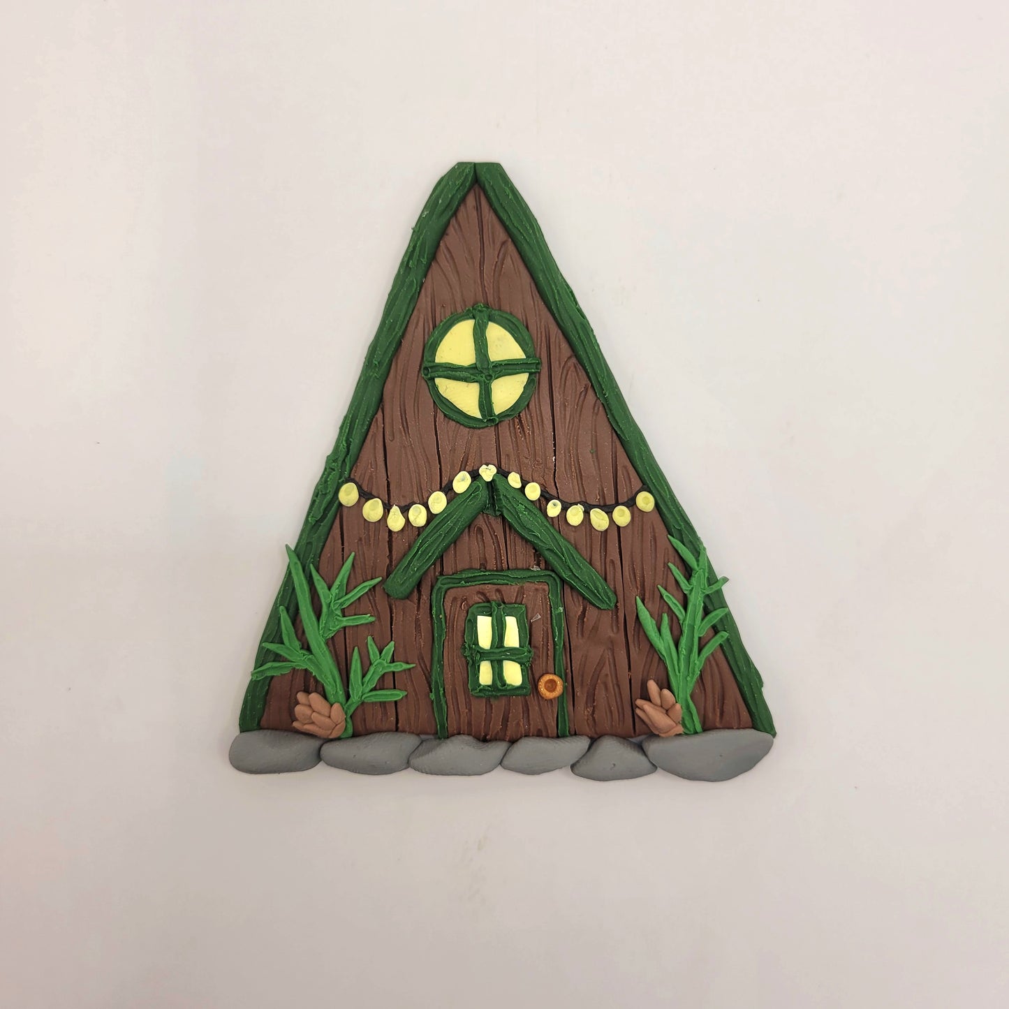 The alpine cabin fairy door rests on a white background. It is brown with a forest green roof and detailed with a window, fairy lights, door, spruce sprigs and pinecones.
