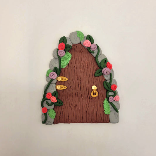 The fairy door rests on a white background. The secret garden door is a maroon wood pattern with grey cobblestone finish and gold hinges and door handle. It is detailed with green vines and pink and purple flowers.