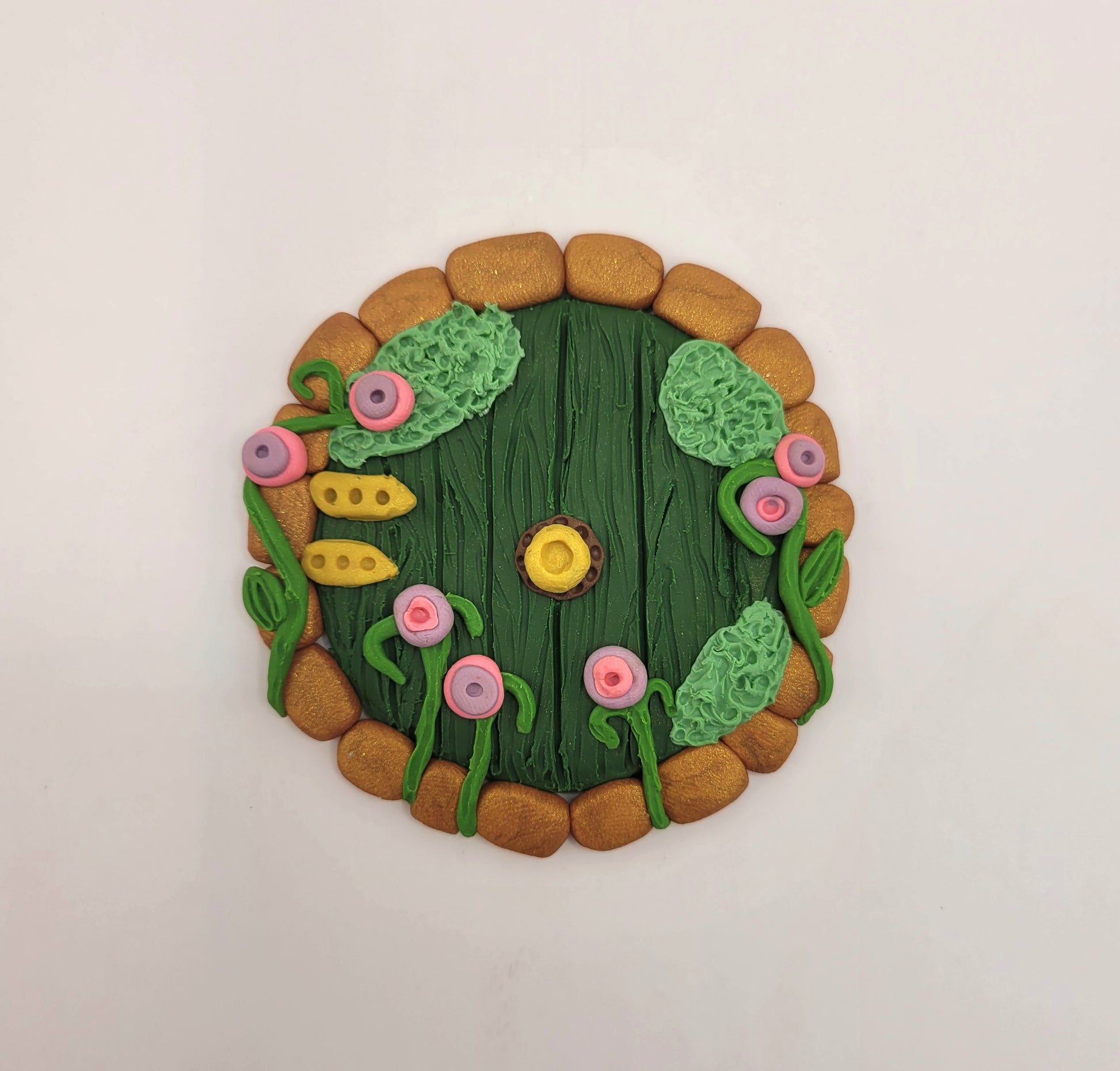 The Hobbit door rests on a white background. It is circular and green wood patterned with bronze brick finishes and golden hinges and a doorknob. It is detailed with green grass, moss and bright purple and pink flowers.