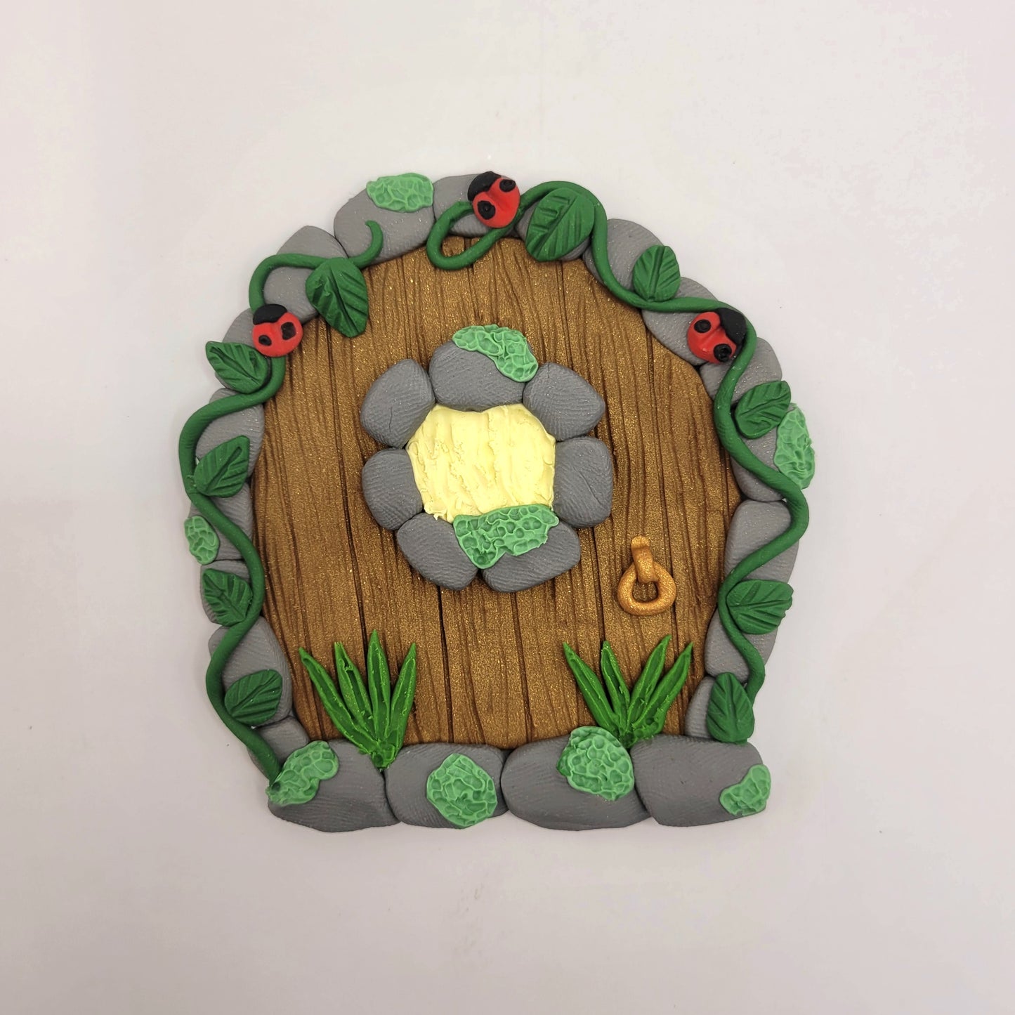 The ladybug fairy door rests on a white background. The mini ladybug door is brown wood patterned with grey cobblestone finish, a window and a door knob. It is detailed with green grass, moss, vines and, of course, ladybugs!