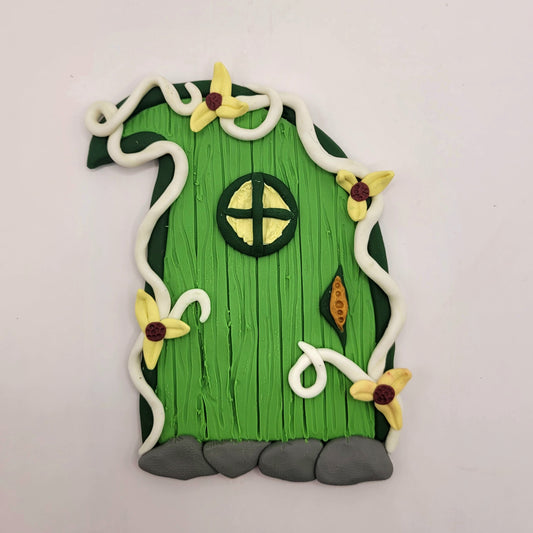 The Elvish fairy door sits on a white background. Shaped like a leaf, this Elvish door has green wood patterning, white vine finish, and a window. It is detailed with yellow flowers.