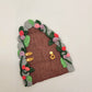 The fairy door is shown in close up at an angle. The secret garden door is a maroon wood pattern with grey cobblestone finish and gold hinges and door handle. It is detailed with green vines and pink and purple flowers.