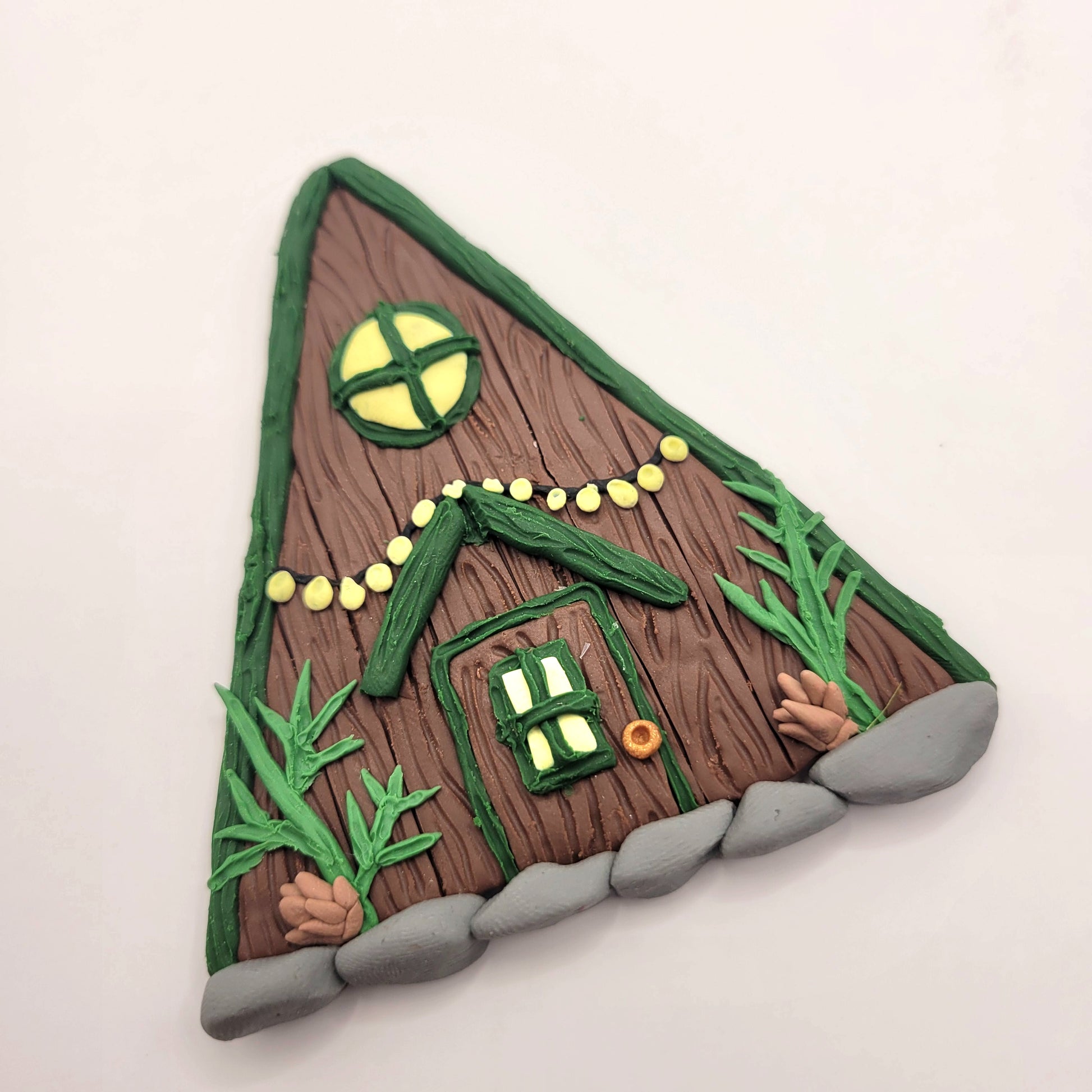 The alpine cabin fairy door rests on a white background at an angle. It is brown with a forest green roof and detailed with a window, fairy lights, door, spruce sprigs and pinecones.