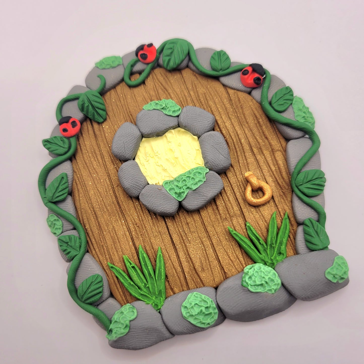 The ladybug fairy door rests at an angle on a white background. The mini ladybug door is brown wood patterned with grey cobblestone finish, a window and a door knob. It is detailed with green grass, moss, vines and, of course, ladybugs!