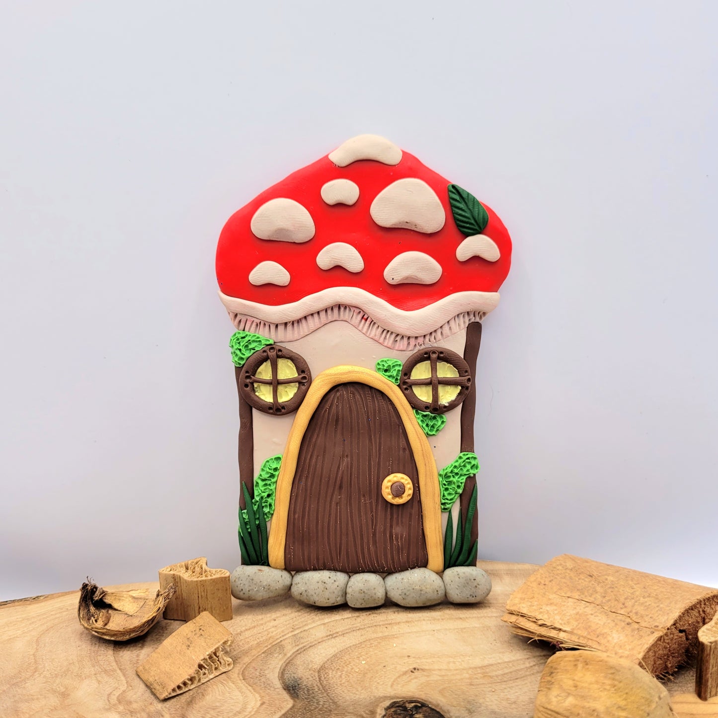The mushroom door rests on an oak wood platform with dried wood pieces. The mushroom's cap is bright red with beige textured spots and a green leaf. The stem of the mushroom is beige and is detailed with two windows, a door, green moss and grass, and a cobblestone step.