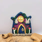 The fairy door rests on an oak wood platform with dried wood pieces. The dark purple door is covered in glitter with a dark blue "wood" roof. It is detailed with gold trim, a window and door, a crystal display, mushrooms, vines and grass.