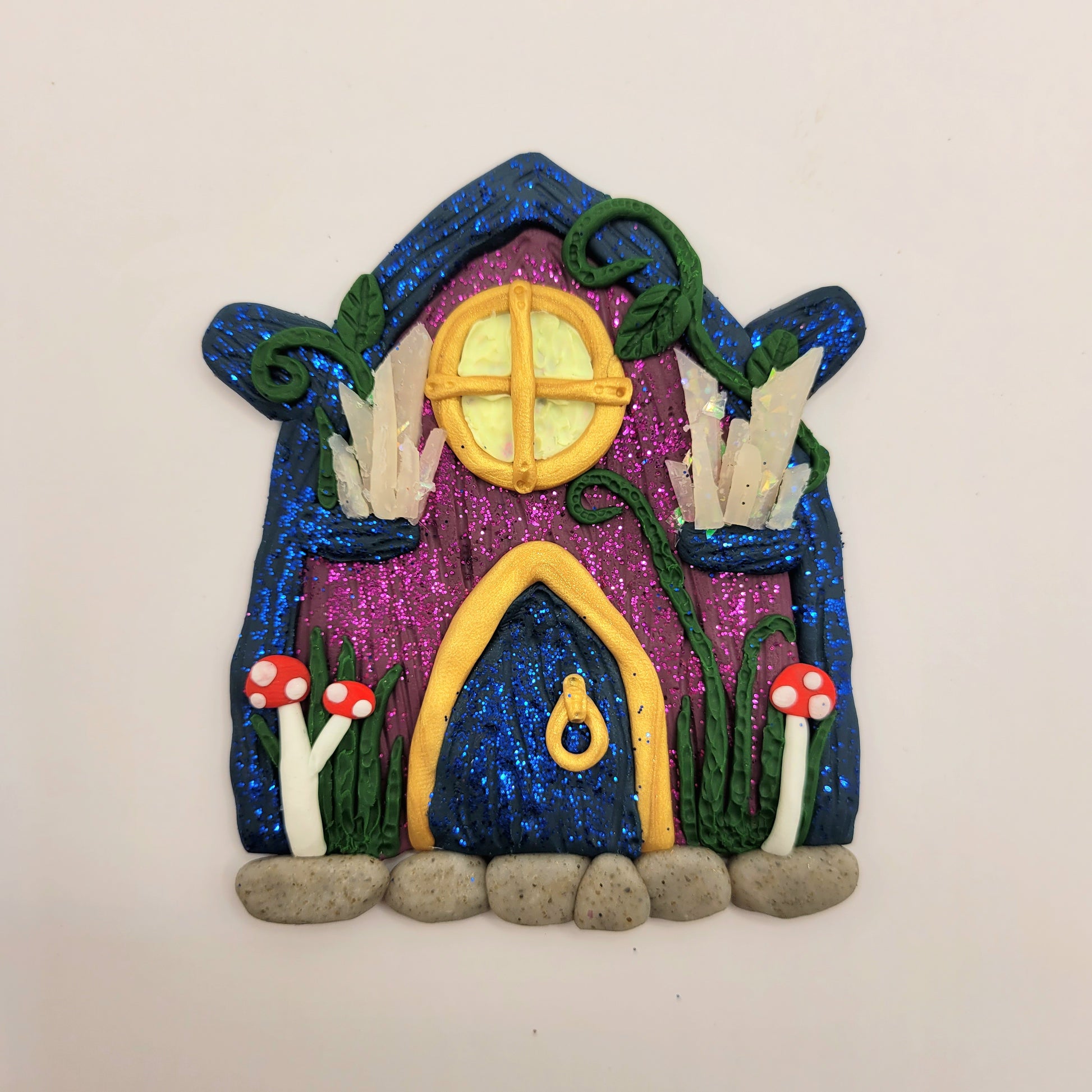 The fairy door is shown against a white background. The dark purple door is covered in glitter with a dark blue "wood" roof. It is detailed with gold trim, a window and door, a crystal display, mushrooms, vines and grass.