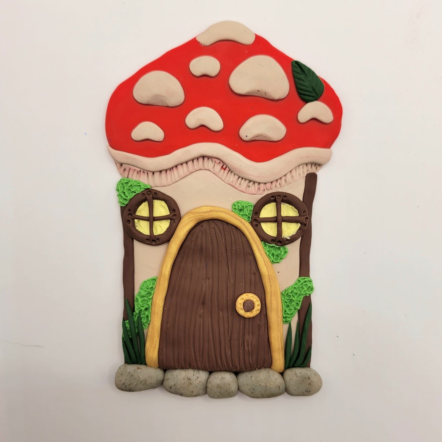 The mushroom door rests on a white background. The mushroom's cap is bright red with beige textured spots and a green leaf. The stem of the mushroom is beige and is detailed with two windows, a door, green moss and grass, and a cobblestone step.