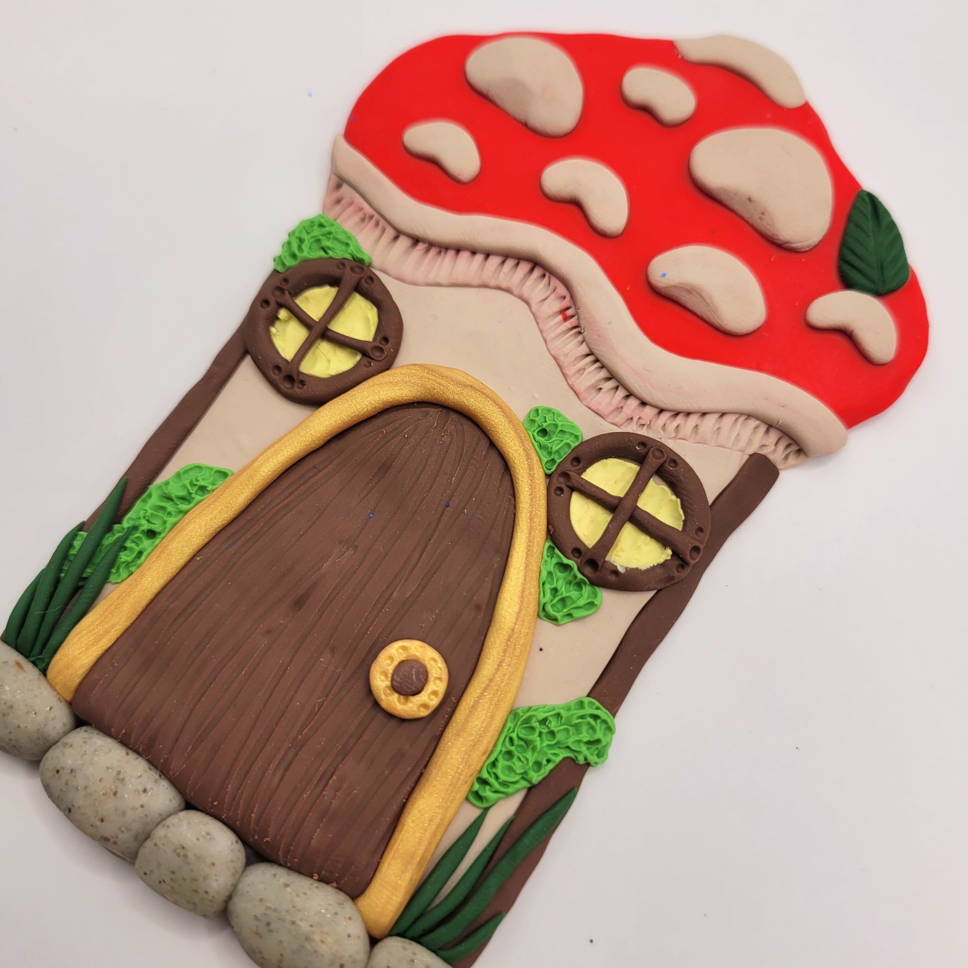 The mushroom door is shown at a close up angle. The mushroom's cap is bright red with beige textured spots and a green leaf. The stem of the mushroom is beige and is detailed with two windows, a door, green moss and grass, and a cobblestone step.