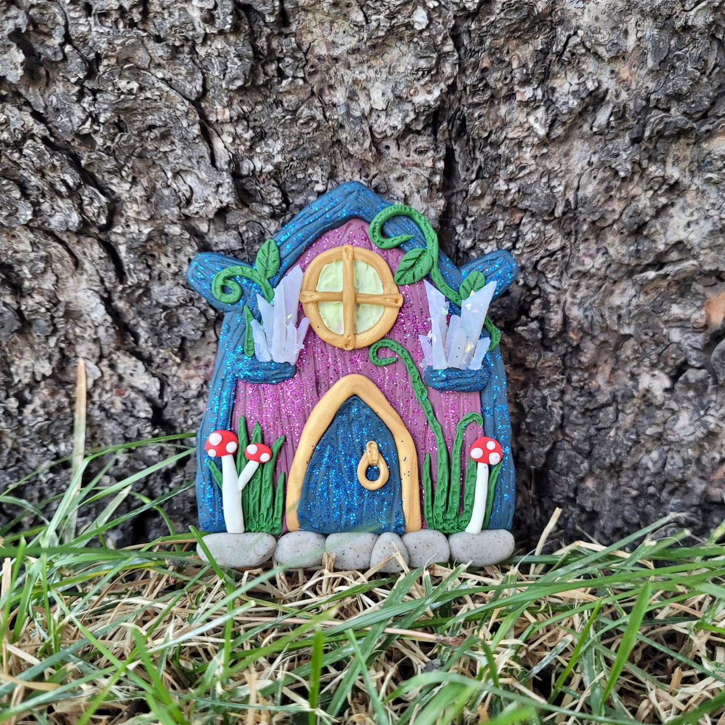 The fairy door rests against the bark of a tree surrounded by green grass. The dark purple door is covered in glitter with a dark blue "wood" roof. It is detailed with gold trim, a window and door, a crystal display, mushrooms, vines and grass.