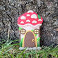 The mushroom door rests against the bark of a tree outside surrounded by green grass. The mushroom's cap is bright red with beige textured spots and a green leaf. The stem of the mushroom is beige and is detailed with two windows, a door, green moss and grass, and a cobblestone step.