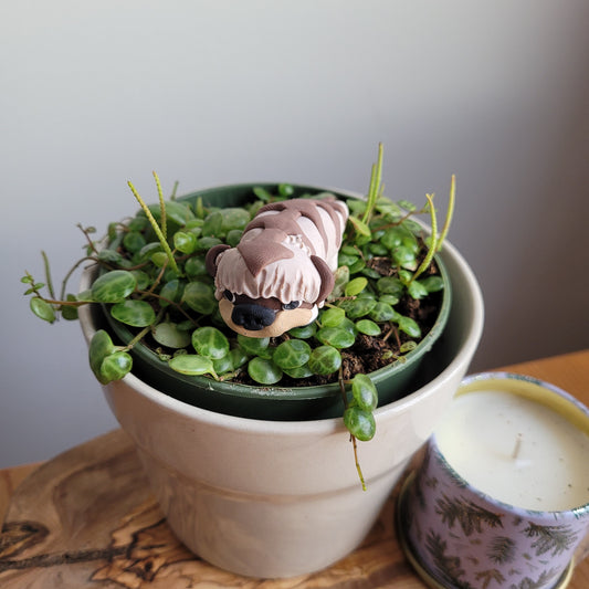 A collectible handmade Appa figurine stands nestled in a houseplant with a white pot next to a candle. The cute Appa figurine is a beige bison with an arrow on its forehead and six legs. 
