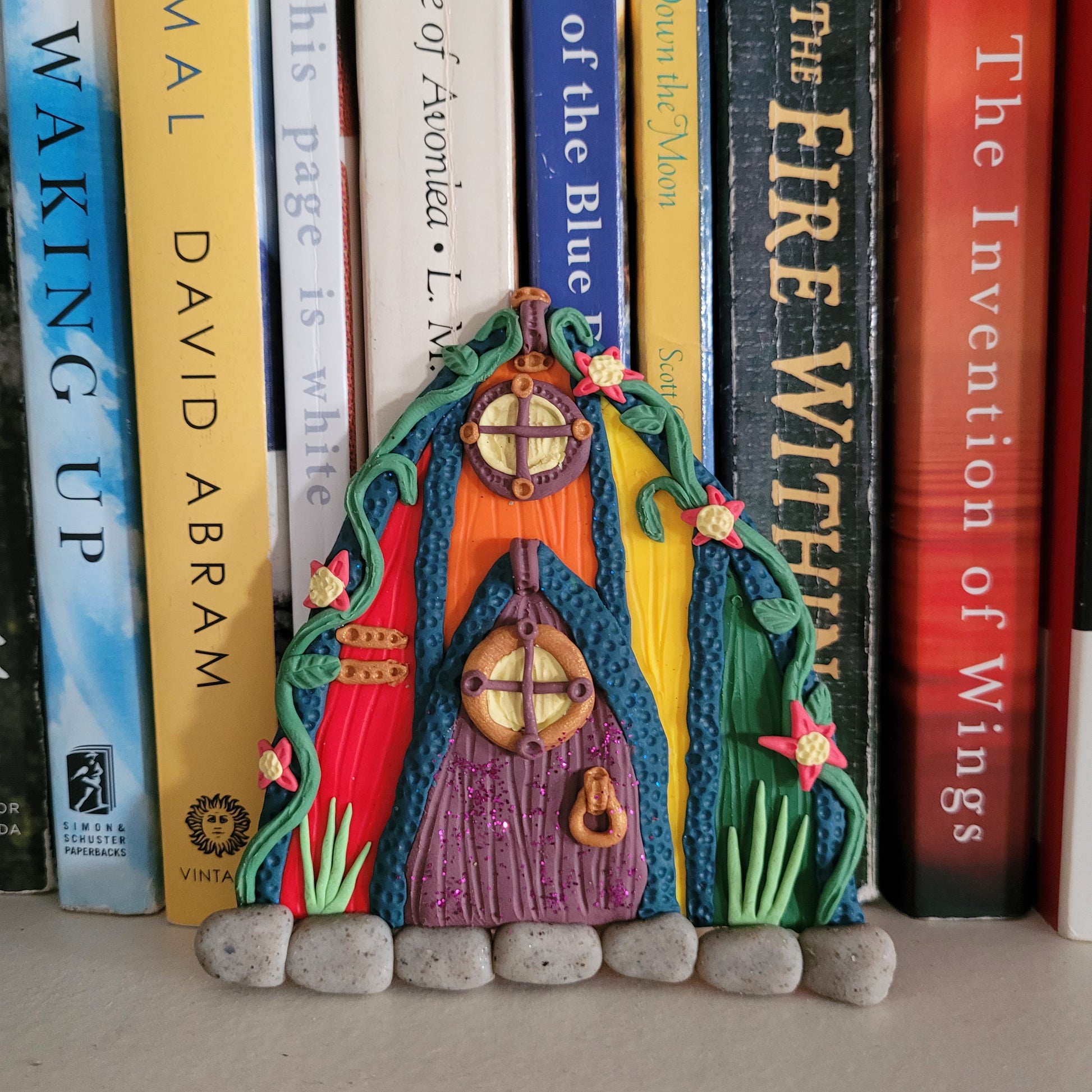 The Pride fairy door is rainbow colored and rests against a shelf of colorful books as the perfect decor.