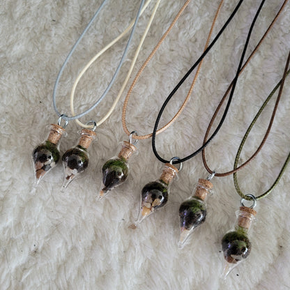 Six tiny terrarium necklaces lie on a white plush rug. The necklace comes in six colours: light blue, beige, light brown, black, brown and forest green. By Planted Glass Terrariums in Edmonton.