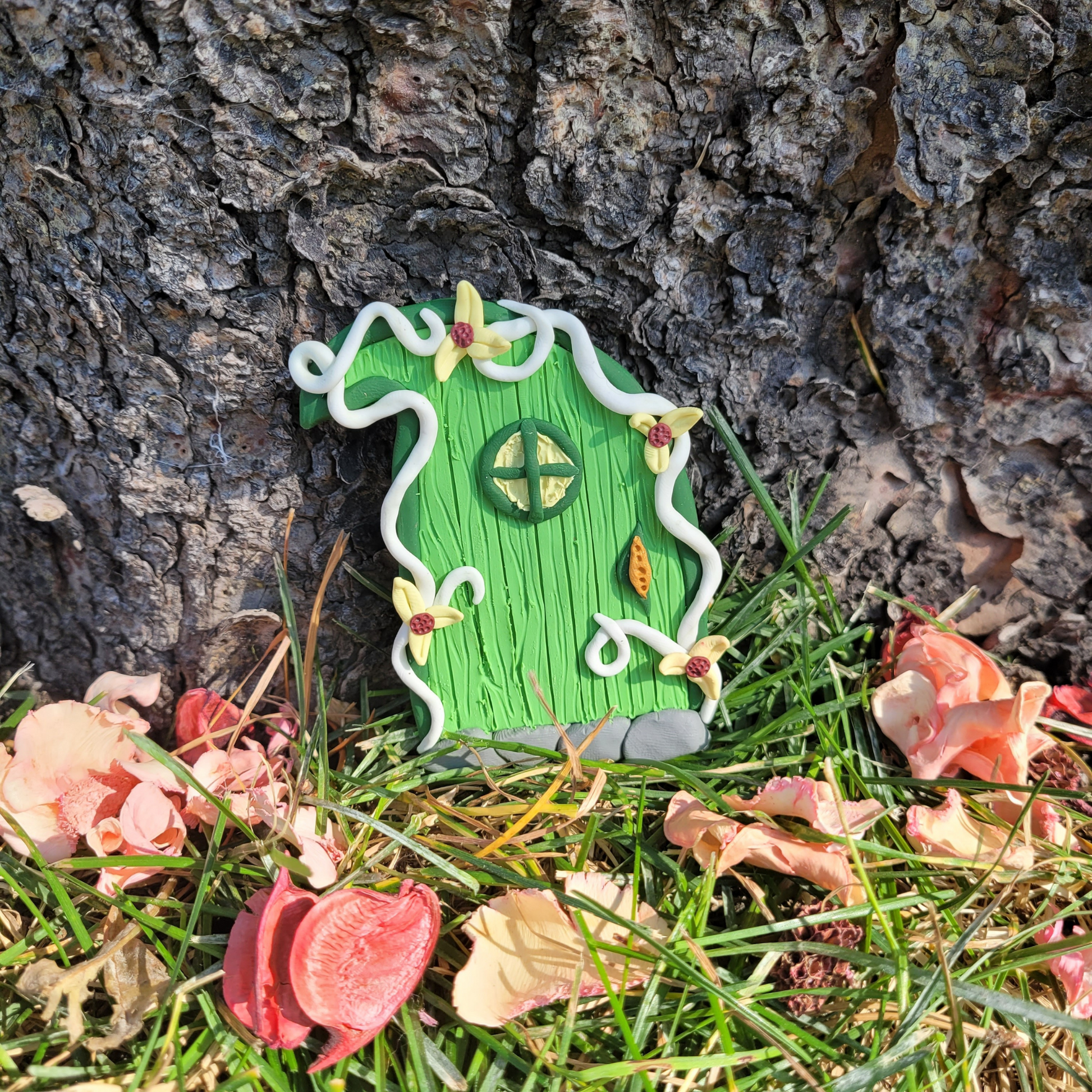 The Elvish fairy door rests against the bark of a tree, surrounded by grass and pink flower petals. Shaped like a leaf, this Elvish door has green wood patterning, white vine finish, and a window. It is detailed with yellow flowers.